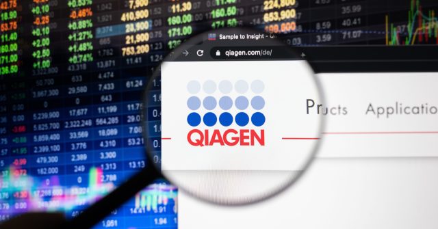 Das Qiagen-Firmenlogo auf einer Website mit verschwommenen Börsenentwicklungen im Hintergrund, gesehen auf einem Computerbildschirm durch ein Vergrößerungsglas
