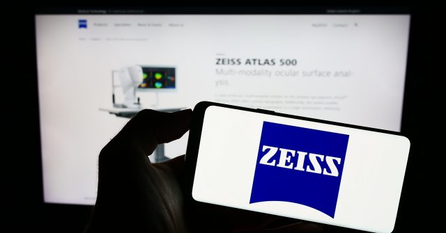 Eine Person hält ein Mobiltelefon mit dem Logo des Medizintechnikunternehmens Carl Zeiss Meditec AG auf dem Bildschirm vor einer Webseite mit Fokus auf das Telefondisplay