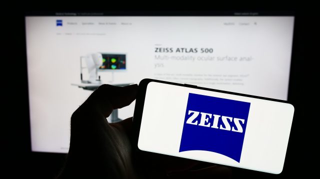 Eine Person hält ein Mobiltelefon mit dem Logo des Medizintechnikunternehmens Carl Zeiss Meditec AG auf dem Bildschirm vor einer Webseite mit Fokus auf das Telefondisplay