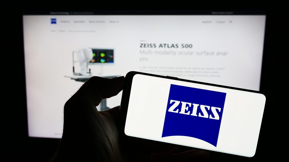 Eine Person hält ein Mobiltelefon mit dem Logo des Medizintechnikunternehmens Carl Zeiss Meditec AG auf dem Bildschirm vor einer Webseite mit Fokus auf das Telefondisplay