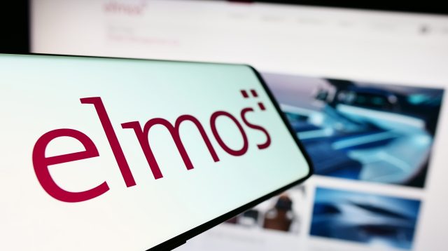Mobiltelefon mit dem Logo der deutschen Firma Elmos Semiconductor SE auf dem Bildschirm vor einer Unternehmenswebsite mit Fokus auf die Mitte links des Telefondisplays