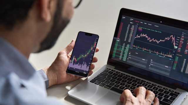 Mann analysiert Aktienkurse auf Handy und Laptop