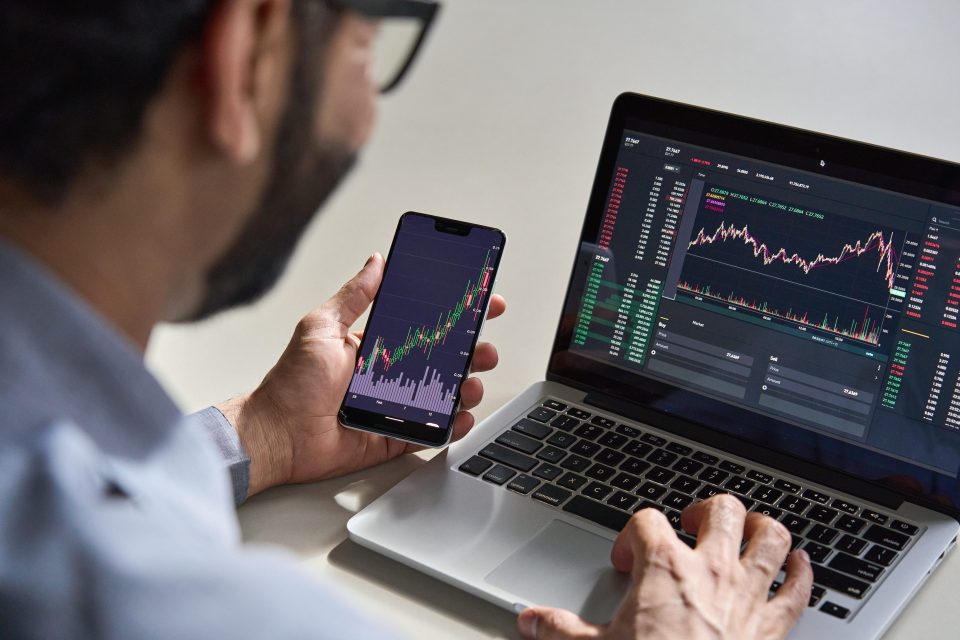 Mann analysiert Aktienkurse auf Handy und Laptop