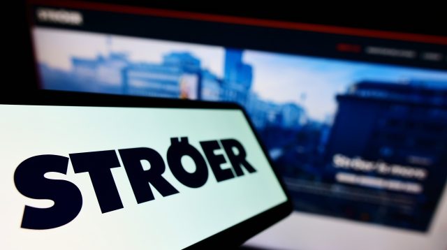 Mobiltelefon mit Logo der deutschen Werbefirma Ströer SE Co. KGaA auf dem Bildschirm vor einer Unternehmenswebsite mit Fokus auf der linken Seite des Telefondisplays