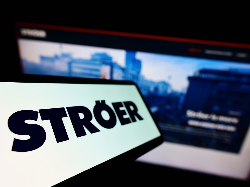 Mobiltelefon mit Logo der deutschen Werbefirma Ströer SE Co. KGaA auf dem Bildschirm vor einer Unternehmenswebsite mit Fokus auf der linken Seite des Telefondisplays