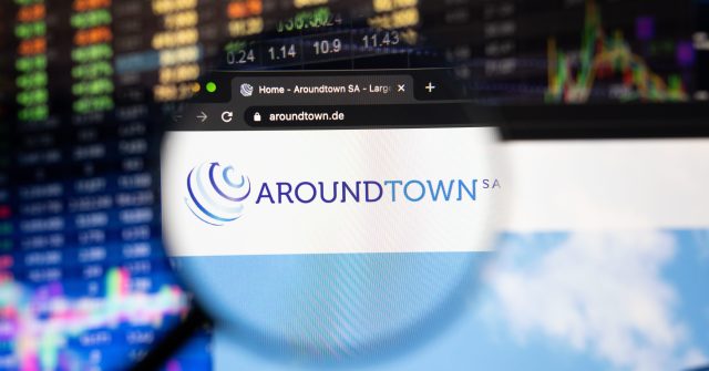 Aroundtown SA Firmenlogo auf einer Website mit verschwommenen Börsenentwicklungen im Hintergrund, gesehen auf einem Computerbildschirm durch eine Lupe