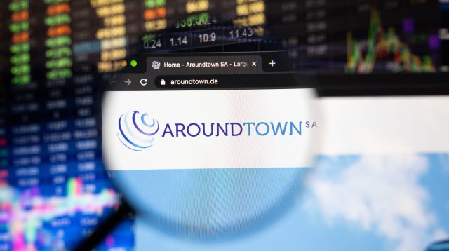 Aroundtown SA Firmenlogo auf einer Website mit verschwommenen Börsenentwicklungen im Hintergrund, gesehen auf einem Computerbildschirm durch eine Lupe