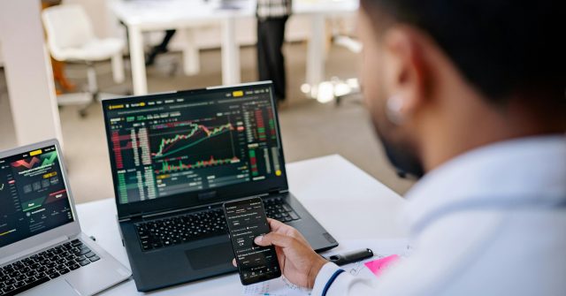 Mann analysiert Aktienkurse auf Handy und Laptop