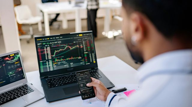 Mann analysiert Aktienkurse auf Handy und Laptop