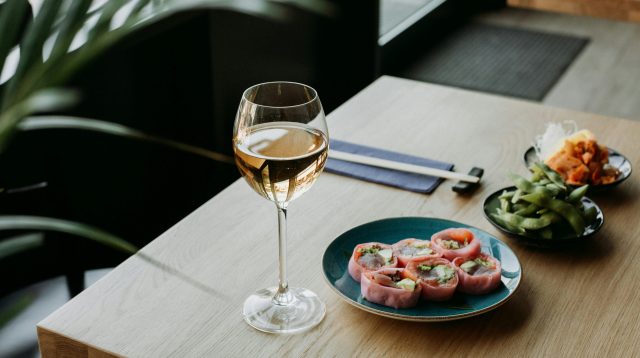Glas Weißwein, Teller mit Sushi und Essstäbchen auf hellem Holztisch